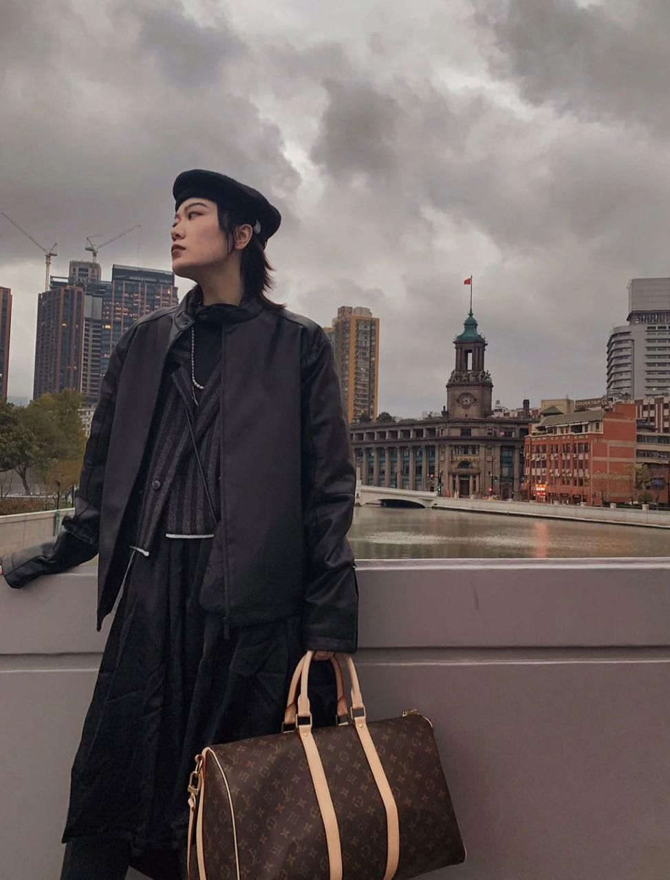 Person in black coat and hat with a brown leather bag against an urban skyline
