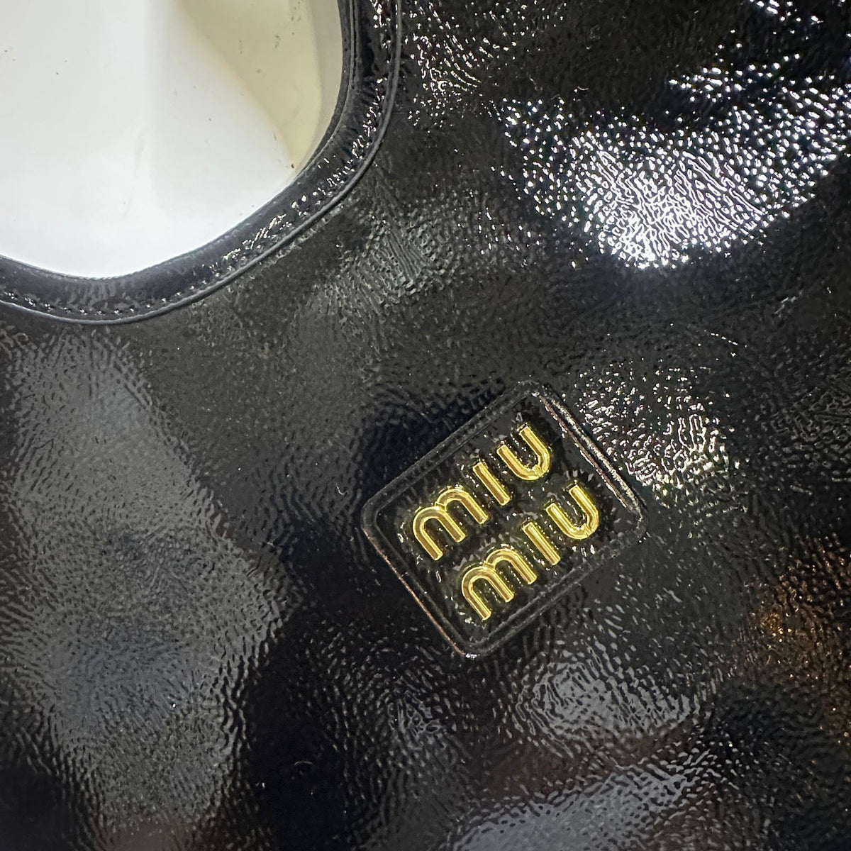 Alt Text: Raised gold metal Miu Miu logo detail on black crinkled glossy leather